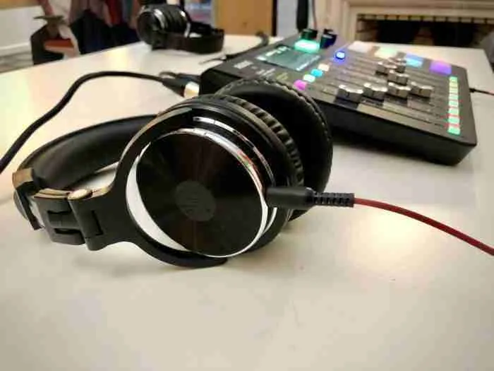 Fone de ouvido preto está em destaque na frente da imagem e está ligado por um cabo a uma mesa de som, que aparece ao fundo um pouco desfocada, com botões coloridos e controles deslizantes.