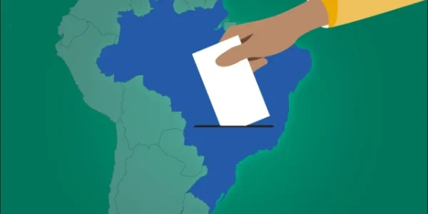 Ilustração de um mapa do Brasil em azul sobre fundo verde, visto de cima. Uma mão parda, vestindo manga amarela, aparece no canto superior direito, colocando um retângulo branco que representa uma cédula de votação sobre a área do mapa, simbolizando o ato de votar no país.