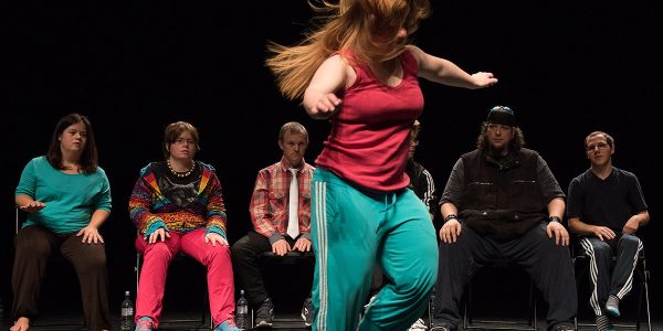 Mulher em cena de dança contemporânea, em palco escuro, aparece em primeiro plano com cabelos longos ruivos em movimento, vestindo regata rosa e calça esportiva verde, enquanto executa um passo com os braços abertos e pés em meia ponta; ao fundo, uma fileira de cinco pessoas sentadas observa a apresentação, com expressões diversas e roupas casuais coloridas, sugerindo um grupo heterogêneo em contexto de teatro inclusivo.