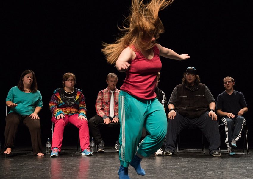 Mulher em cena de dança contemporânea, em palco escuro, aparece em primeiro plano com cabelos longos ruivos em movimento, vestindo regata rosa e calça esportiva verde, enquanto executa um passo com os braços abertos e pés em meia ponta; ao fundo, uma fileira de cinco pessoas sentadas observa a apresentação, com expressões diversas e roupas casuais coloridas, sugerindo um grupo heterogêneo em contexto de teatro inclusivo.