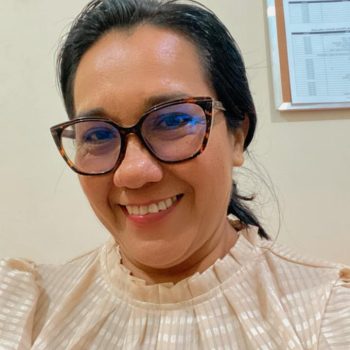 Uma mulher de pele escura, olhos, nariz e boca medianos, cabelos lisos e amarrado, sorri. Usa óculos com uma armação decorada em tons de preto e amarronzado. Veste blusa bege. Ao fundo uma parede clara e o quadro de avisos.