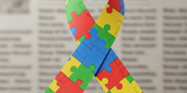 A imagem apresenta um laço colorido formado por peças de quebra-cabeça em tons de azul, vermelho, amarelo e verde. O laço é um símbolo amplamente associado à conscientização sobre o autismo. O fundo da imagem é um jornal desfocado, com textos e colunas visíveis, mas sem nitidez suficiente para leitura. O efeito cria um destaque no laço, que simboliza a diversidade e a complexidade do espectro autista