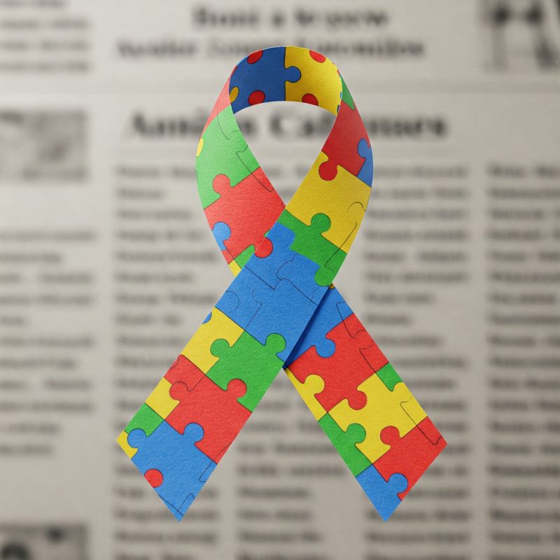 Autismo_Na_Midia A imagem apresenta um laço colorido formado por peças de quebra-cabeça em tons de azul, vermelho, amarelo e verde. O laço é um símbolo amplamente associado à conscientização sobre o autismo. O fundo da imagem é um jornal desfocado, com textos e colunas visíveis, mas sem nitidez suficiente para leitura. O efeito cria um destaque no laço, que simboliza a diversidade e a complexidade do espectro autista