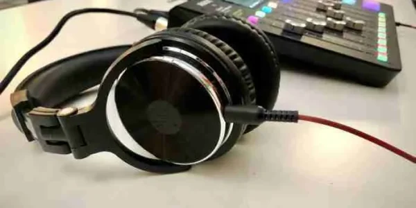 Fone de ouvido preto está em destaque na frente da imagem e está ligado por um cabo a uma mesa de som, que aparece ao fundo um pouco desfocada, com botões coloridos e controles deslizantes.