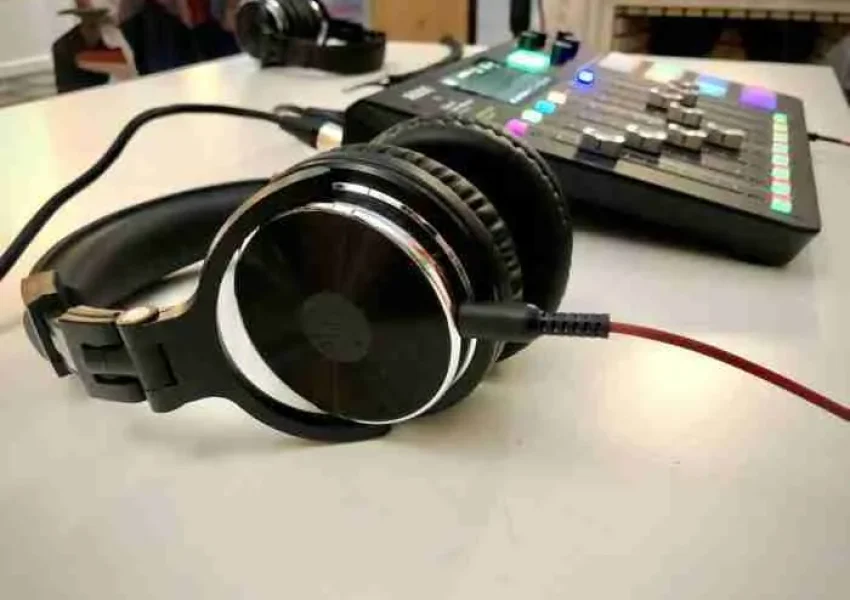 Fone de ouvido preto está em destaque na frente da imagem e está ligado por um cabo a uma mesa de som, que aparece ao fundo um pouco desfocada, com botões coloridos e controles deslizantes.