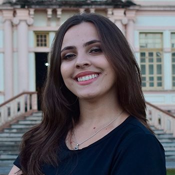 Foto de Isabela da Matta, mulher branca, cabelos lisos e castanhos. Na foto, Isabela mostra os braços cruzados, sorri e veste uma camisa preta com os dizeres: "Formandos/ Universidade Federal de Viçosa/ Julho 2023". Ao fundo, vê-se parte da fachada de um prédio antigo.