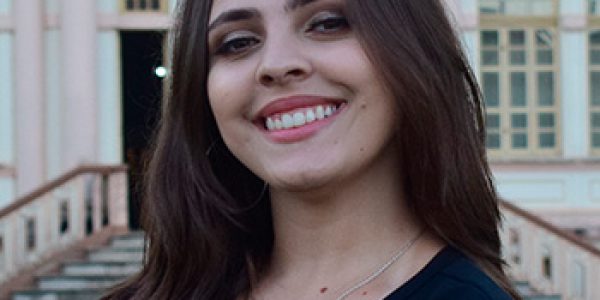 Foto de Isabela da Matta, mulher branca, cabelos lisos e castanhos. Na foto, Isabela mostra os braços cruzados, sorri e veste uma camisa preta com os dizeres: "Formandos/ Universidade Federal de Viçosa/ Julho 2023". Ao fundo, vê-se parte da fachada de um prédio antigo.