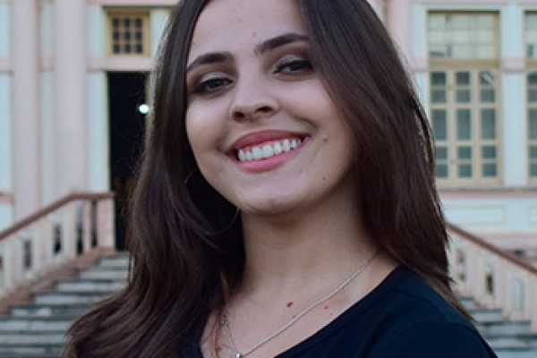 Foto de Isabela da Matta, mulher branca, cabelos lisos e castanhos. Na foto, Isabela mostra os braços cruzados, sorri e veste uma camisa preta com os dizeres: "Formandos/ Universidade Federal de Viçosa/ Julho 2023". Ao fundo, vê-se parte da fachada de um prédio antigo.