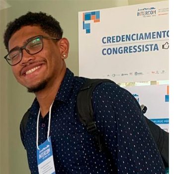 Foto de João Faustino, homem de 1,80 e pele escura, autodeclarado preto, com o cabelo ondulado curto, e de olhos castanhos escuros. Na imagem, ele está sorrindo e utilizando um óculos de armação preta, uma camisa azul escuro, com preenchida com a frase “All boy”. O fundo da foto se refere a um espaço de jardim.