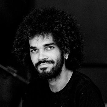 Foto em preto e branco de meio corpo de Marcos Fernandes, um homem de pele parda, cabelo preto e crespo em formato "Black Power". Ele tem uma barba preenchendo todo rosto e preta. Em sua orelha esquerda dois pequenos brincos prateados em formato de argola. Ele está sentado sob um braço de uma cadeira apoiando-se em outra a sua frente. Está usando uma calça moletom preta com pequenos detalhes brancos horizontais formando uma estampa. Usa uma camisa preta com estampa do 6º Festival de Música da Paraíba, que forma-se através de pequenas notas musicais viajando através de uma grande clave de sol. Marcos está sorrindo levemente e o fundo é desfocado, mas é perceptível a sua esquerda um tripé de câmera e a sua direita uma mesa redonda e branca.