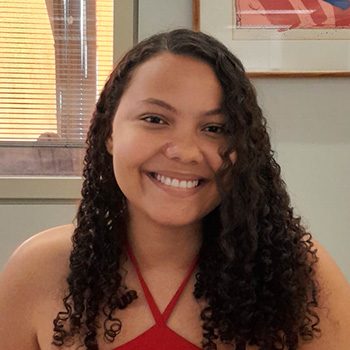 Foto de meio corpo de Rúbia Layane, uma mulher de olhos castanhos e cabelos cacheados que descem um pouco abaixo do ombro. Ela usando uma camiseta vermelha com amarração no pescoço. Rúbia está sorrindo abertamente, o fundo da foto é uma parede cinza, com uma janela ao canto esquerdo e um quadro multicolorido no lado direito.