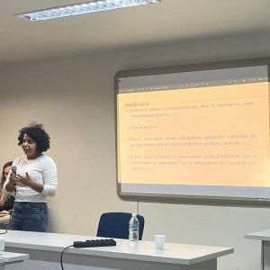 Foto da pesquisadora Talita França, uma mulher parda de cabelos curtos escuros, vestindo blusa branca e calça jeans azul, fala ao microfone, em pé, por trás de um birô. Atrás dela, uma projeção de texto numa tela branca. apresentando sua pesquisa