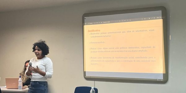 Foto da pesquisadora Talita França, uma mulher parda de cabelos curtos escuros, vestindo blusa branca e calça jeans azul, fala ao microfone, em pé, por trás de um birô. Atrás dela, uma projeção de texto numa tela branca. apresentando sua pesquisa