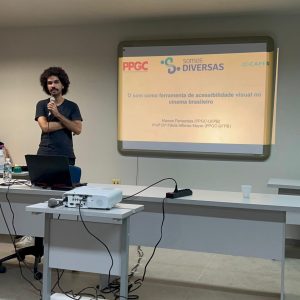 Foto do mestrando Marcos Veloso, um homem negro de pele clara, cabelo black power, vestindo camisa e calça pretas, em pé, segurando um microfone com os braços cruzados. Ele está ao lado de uma tela de projeção e por trás de um birô.