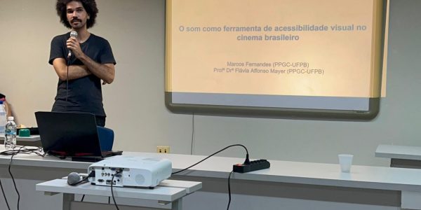 Foto do mestrando Marcos Veloso, um homem negro de pele clara, cabelo black power, vestindo camisa e calça pretas, em pé, segurando um microfone com os braços cruzados. Ele está ao lado de uma tela de projeção e por trás de um birô.