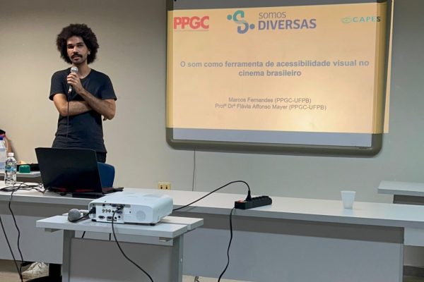 Foto do mestrando Marcos Veloso, um homem negro de pele clara, cabelo black power, vestindo camisa e calça pretas, em pé, segurando um microfone com os braços cruzados. Ele está ao lado de uma tela de projeção e por trás de um birô.