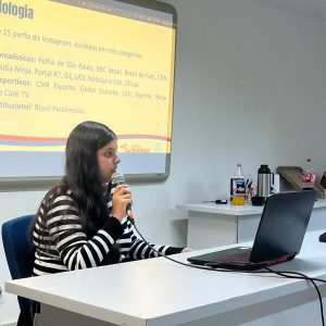 Foto da pesquisadora Thais de Araújo, mulher branca de cabelos escuros e longos. Ela veste uma camisa de mangas compridas de listras, brancas e azuis, e está sentada em um birô, segurando um microfone, e olhando para o notebook à sua frente. Atrás dela, uma tela em branco projeta a apresentação do trabalho.