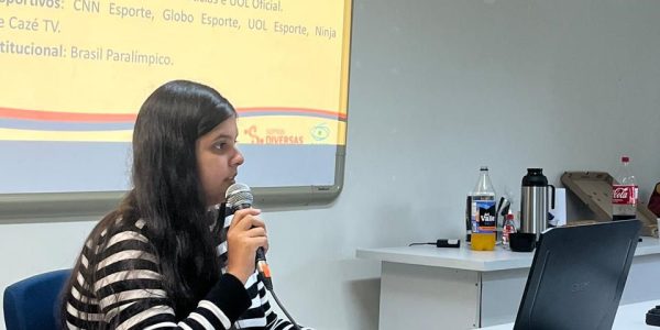 Foto da pesquisadora Thais de Araújo, mulher branca de cabelos escuros e longos. Ela veste uma camisa de mangas compridas de listras, brancas e azuis, e está sentada em um birô, segurando um microfone, e olhando para o notebook à sua frente. Atrás dela, uma tela em branco projeta a apresentação do trabalho.