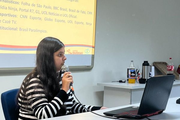 Foto da pesquisadora Thais de Araújo, mulher branca de cabelos escuros e longos. Ela veste uma camisa de mangas compridas de listras, brancas e azuis, e está sentada em um birô, segurando um microfone, e olhando para o notebook à sua frente. Atrás dela, uma tela em branco projeta a apresentação do trabalho.