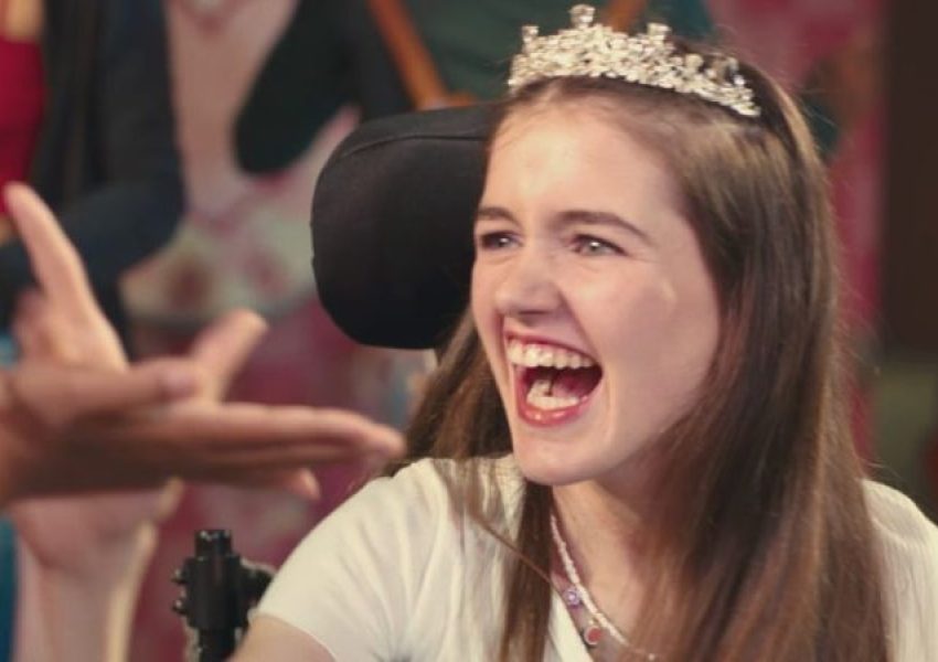 Foto mostra jovem branca de cabelos longos e castanhos, sentada em uma cadeira de rodas com apoio de cabeça. Ela usa uma tiara prateada e sorri com a boca aberta, enquanto olha para o lado. Ao fundo, aparecem outras pessoas desfocadas, em um ambiente colorido.
