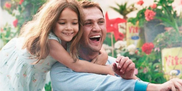 Foto mostra uma menina branca sorridente abra&ccedil;ando um homem branco por tr&aacute;s, com os bra&ccedil;os ao redor do seu pesco&ccedil;o. Ele tamb&eacute;m sorri e olha para frente. Ao fundo, uma vegeta&ccedil;&atilde;o com flores e objetos coloridos.