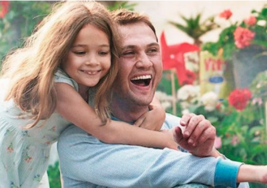 Foto mostra uma menina branca sorridente abraçando um homem branco por trás, com os braços ao redor do seu pescoço. Ele também sorri e olha para frente. Ao fundo, uma vegetação com flores e objetos coloridos.