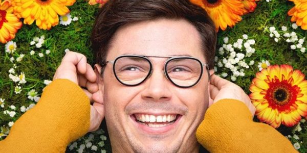 Homem branco jovem com óculos de armação redonda sorri, está deitado de costas sobre um fundo preenchido por flores coloridas e folhagens verdes, vestindo um casaco amarelo mostarda que enquadra seu rosto.