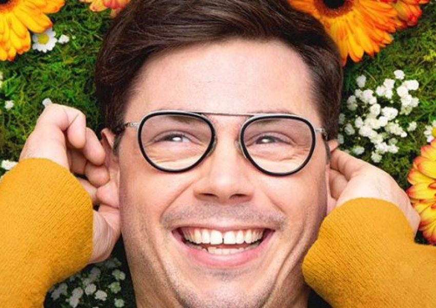 Homem branco jovem com óculos de armação redonda sorri, está deitado de costas sobre um fundo preenchido por flores coloridas e folhagens verdes, vestindo um casaco amarelo mostarda que enquadra seu rosto.